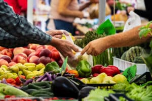 a buyer and a vendor exchange fresh vegetables at a busy farmers market. colorful produce including peppers, peaches, eggplants, asparagus, and other seasonal items fills the foreground. the scene captures direct trade, local agriculture, and vibrant market activity. ideal for themes related to fresh food, farming, shopping, community markets, organic produce, and healthy eating.